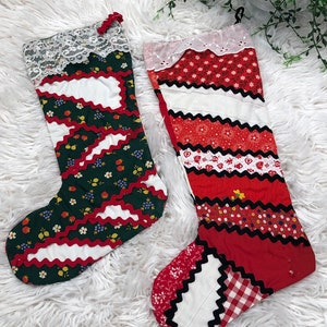 May include: Two fabric stockings featuring patchwork designs. The left stocking is dark green with floral patterns, white sections, and red rickrack trim, topped with white lace. The right stocking is red with various patterns, white sections, and black rickrack trim, topped with white eyelet lace.