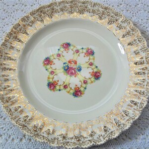 May include: A white ceramic dinner plate with a gold rim and a floral design in the center. The floral design features pink, yellow, blue, and orange flowers.