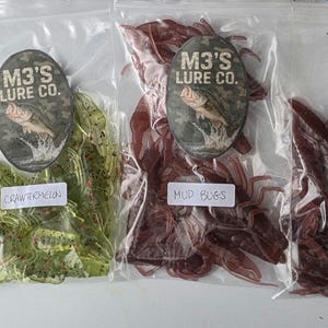 May include: Assortment of soft plastic fishing lures in clear plastic bags. Each bag has a label with "M3'S LURE CO." and a color name. Colors include black, blue, green, red, and brown. The lures are designed to mimic insects.