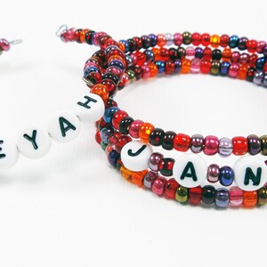 Puede incluir: Dos pulseras de cuentas con los nombres "Aleyah" y "Jane" deletreados en cuentas blancas con letras negras. Las pulseras están hechas con cuentas multicolores en tonos rojos, naranjas, marrones y morados.