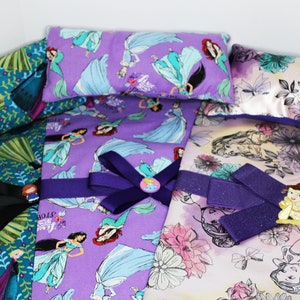 May include: Four purple and white fabric doll beds with princess themed designs. Each bed has a matching pillow with a princess design. The beds have purple ribbon bows and a princess charm.