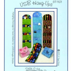 May include: A light blue background with a white frame containing several fabric USB hang-up wallets in various colors and patterns. The wallets are green, blue, and brown with polka dots and other designs. The text "USB Hang-Ups" and "Susie C Shore Designs" are visible.