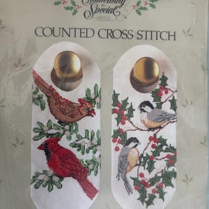 May include: Counted cross stitch door knob decor set featuring chickadees and cardinals. The set includes two white fabric pieces, each with a gold-colored knob at the top. One piece features a red cardinal, the other has chickadees. The text "Something Special" is at the top.