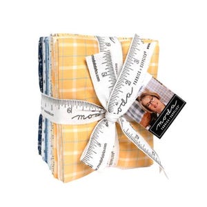 May include: A stack of fabric squares in shades of yellow and blue, tied with a white ribbon printed with a ruler design and the text "moda fabrics". A tag with a woman's photo and the words "moda fabrics + supplies" is attached.