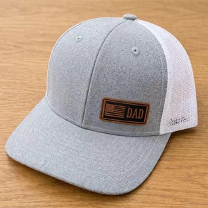 May include: Grey baseball cap with a white mesh back. The cap has a brown leather patch displaying an American flag and the word "DAD". A fashionable accessory for casual wear.