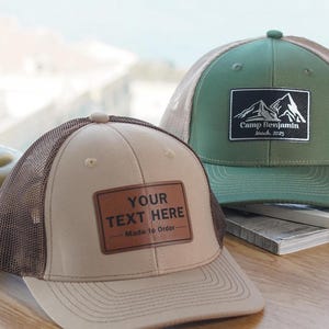 Pode incluir: Dois bonés de caminhoneiro, um bege com malha marrom e um remendo de couro marrom que diz "YOUR TEXT HERE - Made to Order", e outro verde com malha bege e um remendo preto e branco que diz "Camp Benjamin - March 2023".