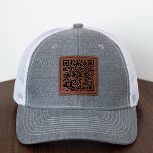 Könnte beinhalten: Graue Trucker-Mütze mit weißem Mesh-Rücken und einem braunen quadratischen Aufnäher mit einem QR-Code. Die Mütze hat einen gebogenen Schirm und einen Knopf oben.
