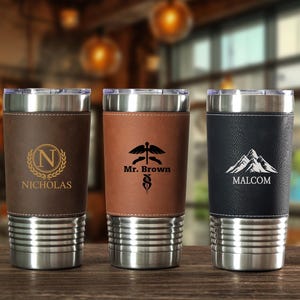May include: Three stainless steel insulated tumblers with leather sleeves. The first tumbler has a brown leather sleeve with the name "Nicholas" and a gold "N" logo. The second has a brown sleeve with "Mr. Brown" and a medical symbol. The third has a black sleeve with "Malcom" and a mountain logo.