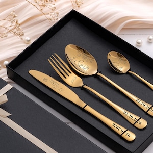 May include: A gold-colored flatware set, including a knife, fork, and two spoons, presented in a black gift box. The utensils are engraved with initials and a date. The set is displayed with a ribbon and a champagne glass.