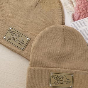 Può includere: Due berretti di maglia beige. Il berretto più grande ha una toppa rettangolare con le parole "Brian", "Frank" e "Daddy" attorno a una grafica di pugno. Il berretto più piccolo ha una toppa simile. I berretti sono su una superficie chiara.