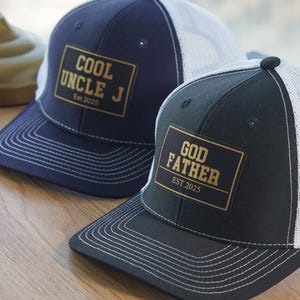 Könnte beinhalten: Zwei schwarz-weiße Trucker-Caps mit goldenen Patches. Die erste Kappe hat eine marineblaue Krone und ein weißes Netzrücken mit einem goldenen Patch, auf dem "COOL UNCLE J Est. 2023" steht. Die zweite Kappe hat eine schwarze Krone und ein weißes Netzrücken mit einem goldenen Patch, auf dem "GOD FATHER EST. 2025" steht.