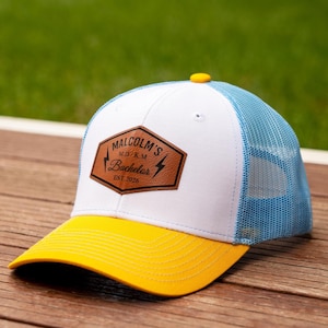 May include: A baseball cap with a bright yellow brim, a white front panel, and a light blue mesh back. A brown faux leather patch on the front features the text MALCOLM'S M.D./K.M. Bachelor EST.2026 with two lightning bolt icons on the sides.