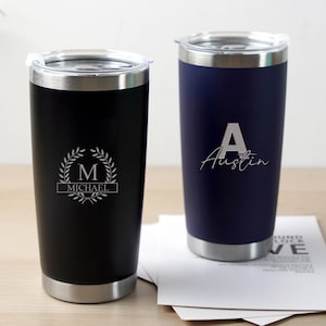 May include: Two personalised stainless steel tumblers. One is black with a silver rim and the name "Michael" below a laurel wreath. The other is navy blue with the name "Austin" in white script. Both have clear lids.