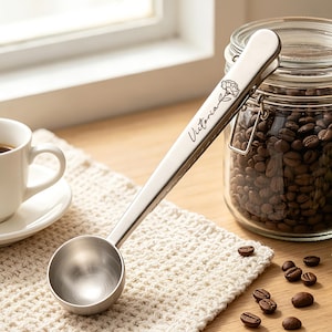 May include: A silver-toned metal coffee scoop with a clip handle featuring the engraved name Victoria and a small floral illustration. The scoop is positioned next to a clear glass jar filled with dark roasted coffee beans.