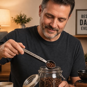 May include: A silver metal coffee scoop with the engraved text "YOU BEAN SO MUCH TO ME, DAD" held over a clear glass jar filled with roasted coffee beans. The jar has a metal wire bail closure and a glass lid with a white rubber seal.