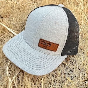 Może przedstawiać: Szara czapka bejsbolowa z czarnym tyłem z siatki. Czapka posiada brązową skórzaną naszywkę z napisem "COACH JACK" i grafiką baseballową. Czapka leży na posłaniu z suchej trawy.