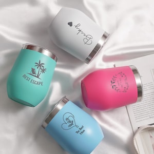 Puede incluir: Cuatro vasos de vino de acero inoxidable con diferentes diseños y colores. Los vasos están sobre una superficie de satén blanco. Los diseños incluyen un corazón con el texto "The Best Mom", una palmera con el texto "Best Escape", una corona floral con el texto "To the Moon and Back", y un corazón con el texto "Boogie".