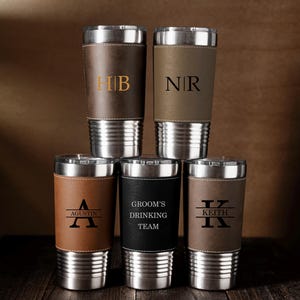 May include: Six personalised travel tumblers with stainless steel bases and lids. The tumblers have leather sleeves in various shades of brown and black. Some are engraved with initials, names, and the text "Groom's Drinking Team".