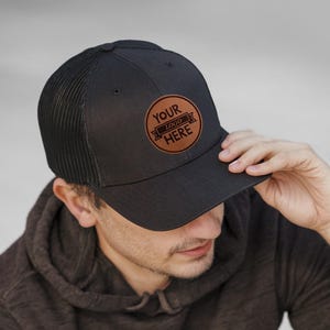 May include: A black and brown trucker hat with a leather patch that says "Your Logo Here".