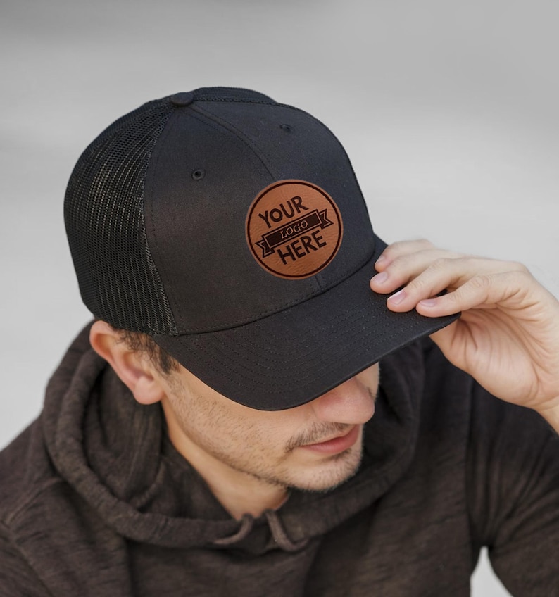 May include: A black and brown trucker hat with a leather patch that says "Your Logo Here".