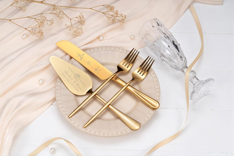 Op de afbeelding: Een goudkleurige bruidstaartmes- en serveerset met gegraveerde namen en een hartontwerp, samen met twee gouden vorken, een champagnefluit en een decoratieve bord. De set is gerangschikt op een wit oppervlak met parels en lint.