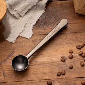May include: A metallic coffee scoop with a long handle featuring the engraved name Hannah and a small flower illustration. The scoop has a deep, rounded bowl and a clip integrated into the handle, finished in a dark, brushed gunmetal tone.