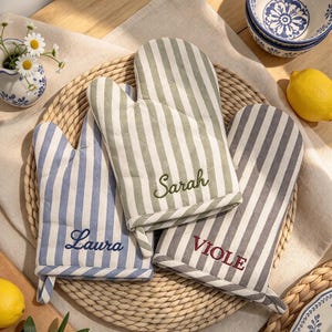 May include: Three oven mitts featuring vertical stripes in sage green, light blue, and grey. Each mitt is embroidered with a name: Sarah in green, Laura in blue, and VIOLE in red. The mitts have a quilted texture and a small fabric loop.