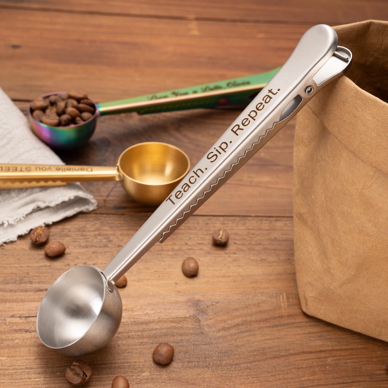 May include: A silver-toned metal coffee scoop with an integrated bag clip handle. The handle features the engraved text "Teach. Sip. Repeat." The scoop has a rounded bowl and a serrated edge on the clip mechanism for a secure grip on bags.