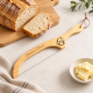 May include: A light-colored wooden bread bow knife with a thin metal wire for slicing. The handle features the engraved text "Gemma's Bread" and a monogram with the letter "G" and the name "Gemma". A brown leather cord is attached to the end.