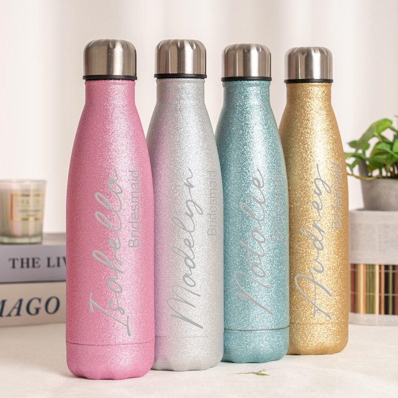 May include: Four glitter-covered water bottles in pink, silver, teal, and gold. Each bottle has a silver lid and a name printed in silver script: Isabella, Madelyn, Natalie, and Audrey. The word "Bridesmaid" is printed below each name.