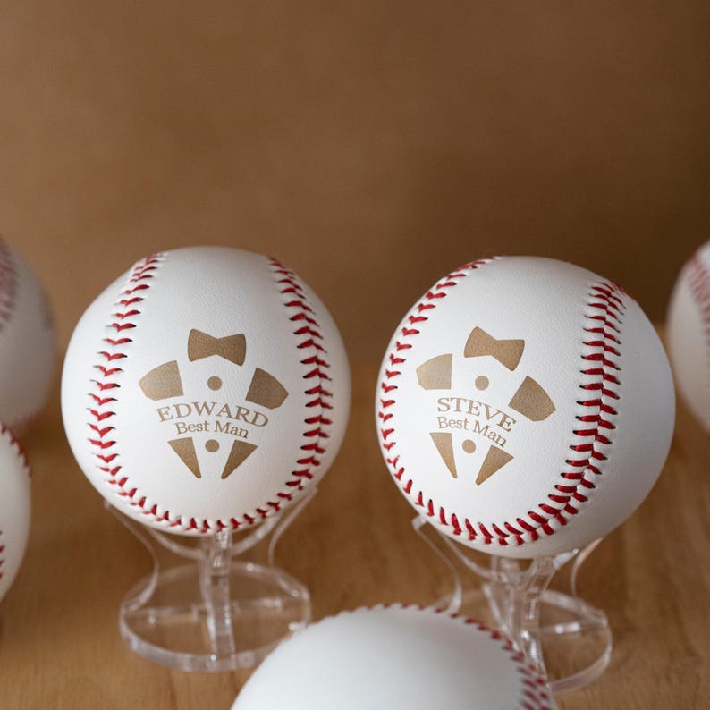 May include: White baseballs with red stitching and personalized text. Each ball features a gold-colored graphic of a tuxedo and the name "Edward Best Man" or "Steve Best Man". The baseballs are displayed on clear stands.