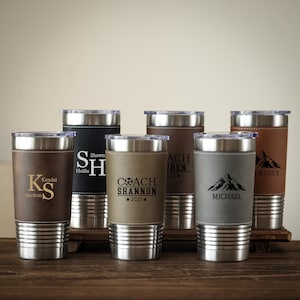 May include: A collection of personalized stainless steel tumblers with leather sleeves. Each tumbler features a different name or initials, with some including text like "COACH SHANNON 2025" and mountain graphics. The tumblers are in various shades of brown, black, and gray.