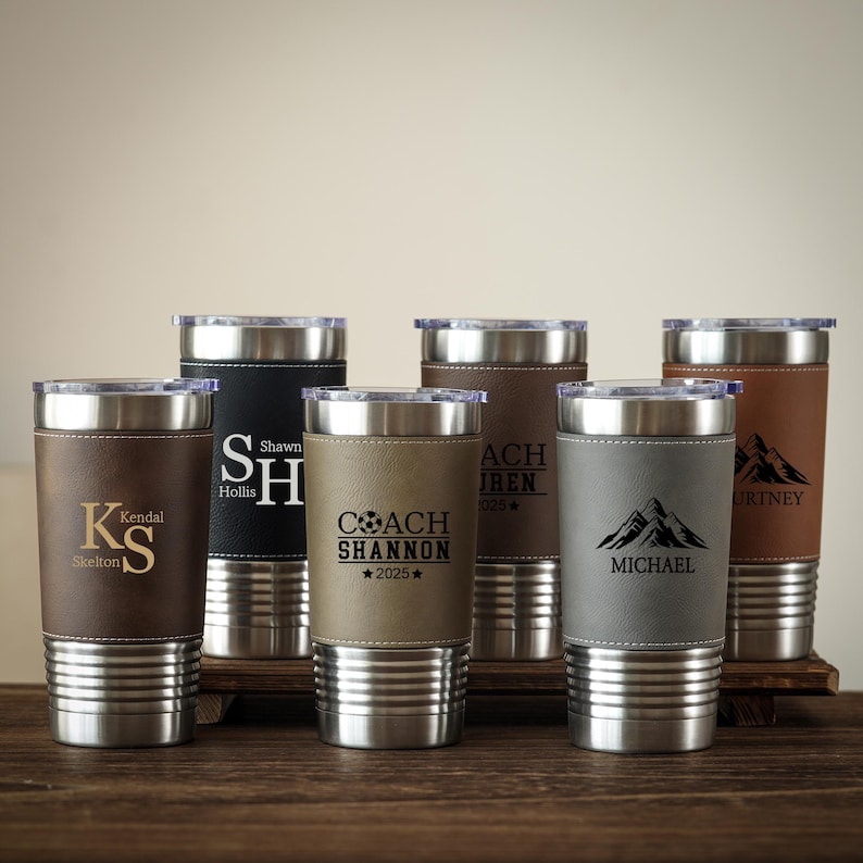 May include: A collection of personalized stainless steel tumblers with leather sleeves. Each tumbler features a different name or initials, with some including text like "COACH SHANNON 2025" and mountain graphics. The tumblers are in various shades of brown, black, and gray.
