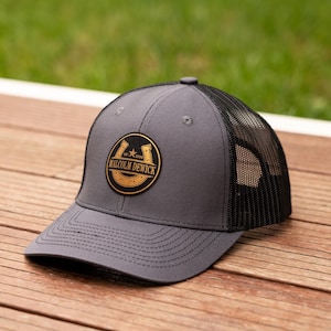 May include: A charcoal grey trucker hat with a black mesh back and a circular patch on the front. The patch features a gold horseshoe design with a star and the text MALCOLM DEWICK EST. 2026. The hat has a structured crown and a curved brim.