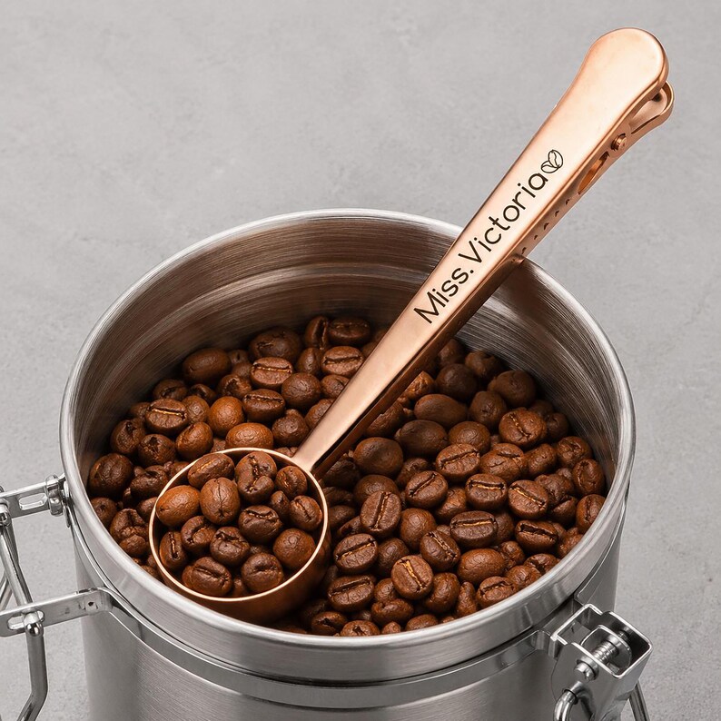 May include: A rose gold coffee scoop with a built-in bag clip, featuring the engraved text "Miss. Victoria" followed by a small coffee bean icon. The scoop is filled with roasted brown coffee beans and rests inside a stainless steel canister.