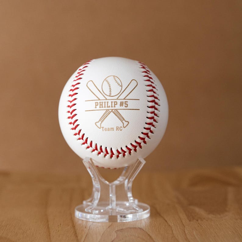 May include: A white baseball with red stitching, displayed on a clear stand. The ball features the text "PHILIP #5" and a baseball bat and ball graphic. The words "Team RC" are also visible.