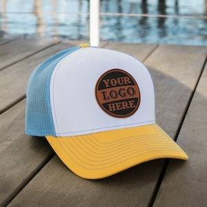 Op de afbeelding: Een witte en blauwe trucker-pet met een gele klep. De pet heeft een bruin leren patch met de tekst "YOUR LOGO HERE" in wit.