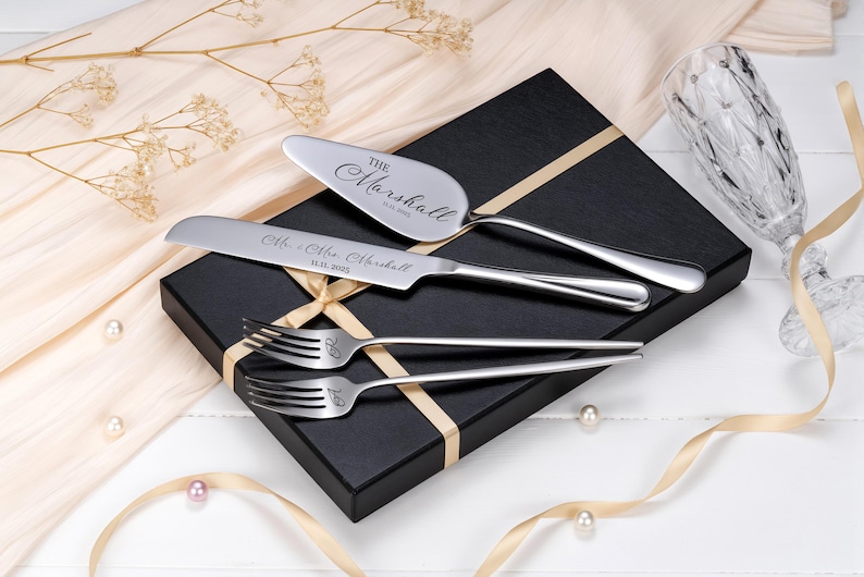 Op de afbeelding: Een zilveren bruidstaartservies, bestaande uit een taartschep, mes en twee vorken, alle gegraveerd met namen en een datum. De set ligt op een zwarte geschenkdoos met een gouden lint. Parels en een champagneglas zijn ook zichtbaar.