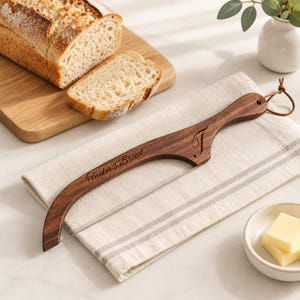 May include: A dark wood bread knife with a thin metal wire blade, featuring the engraved text "Taylor's Bread" and a decorative "T" on the handle. The handle includes a brown leather loop for hanging, showcasing a natural wood grain finish.