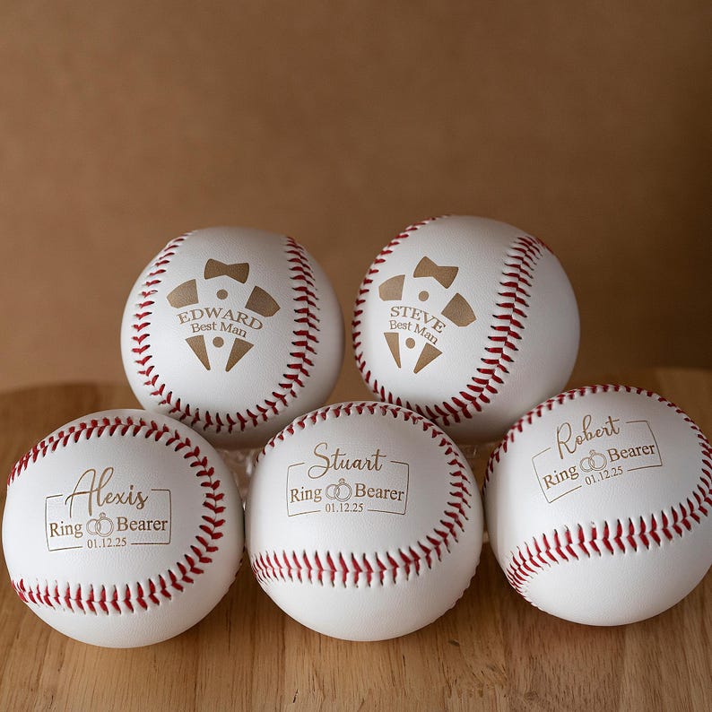 May include: Five white baseballs with red stitching. Each ball is personalized with names and roles like "Best Man" and "Ring Bearer", along with a date. The text is in a gold color, and the baseballs are arranged on a wooden surface.