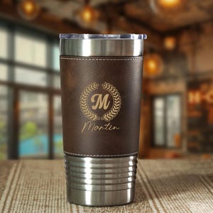May include: A stainless steel travel tumbler with a brown leather wrap. The wrap is laser-etched with a laurel wreath, the letter "M", and the name "Martin". The tumbler has a clear plastic lid.
