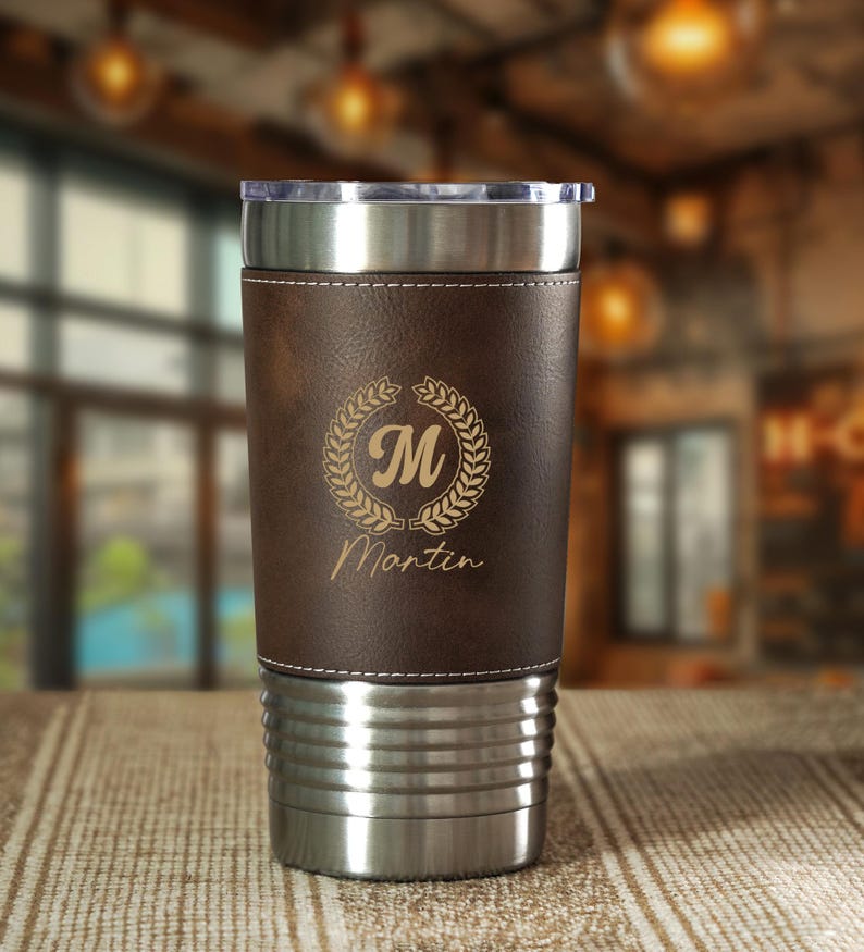 May include: A stainless steel travel tumbler with a brown leather wrap. The wrap is laser-etched with a laurel wreath, the letter "M", and the name "Martin". The tumbler has a clear plastic lid.