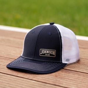 Pode incluir: Um boné trucker azul-marinho com rede branca na parte de trás e aba reta. O painel frontal apresenta um emblema retangular com o texto JOHNSON ESTD.2026 em letras brancas. O boné possui detalhes de costura branca ao longo da aba e coroa.