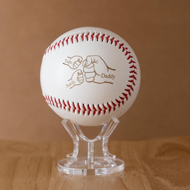 May include: A white baseball with red stitching, displayed on a clear stand. The baseball features an engraved design of three fists, with the words "Tom", "Justin", and "Daddy" inscribed.