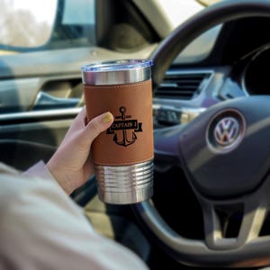 May include: A stainless steel travel mug with a brown leather sleeve. The sleeve features a black anchor design with the text "CAPTAIN J". The mug is held in a car, with the steering wheel and dashboard visible in the background.