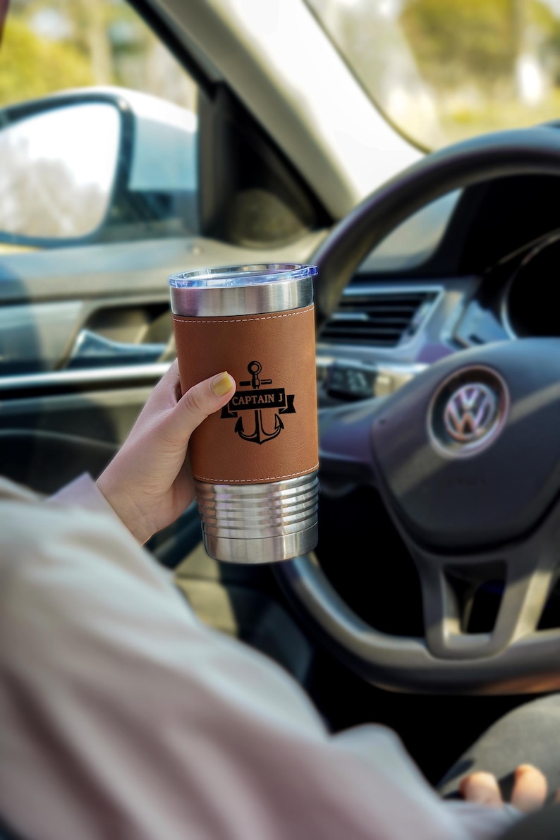 May include: A stainless steel travel mug with a brown leather sleeve. The sleeve features a black anchor design with the text "CAPTAIN J". The mug is held in a car, with the steering wheel and dashboard visible in the background.