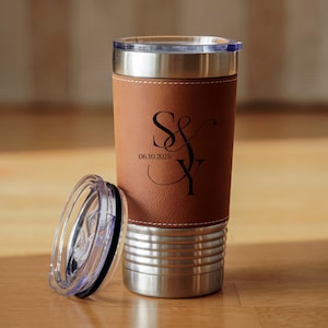 May include: A personalized travel tumbler with a brown leatherette sleeve and a stainless steel base. The tumbler features the initials "S&Y" and the date "06.10.2025" in black script. The clear lid is off to the side.