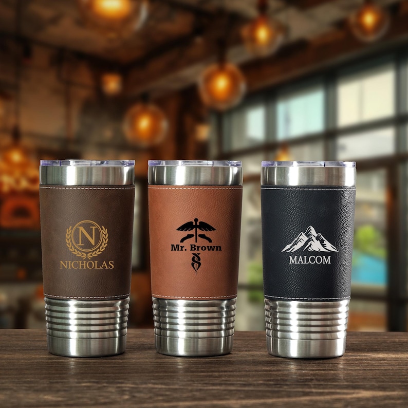 May include: Three stainless steel insulated tumblers with leather sleeves. The first tumbler has a brown leather sleeve with the name "Nicholas" and a gold "N" logo. The second has a brown sleeve with "Mr. Brown" and a medical symbol. The third has a black sleeve with "Malcom" and a mountain logo.
