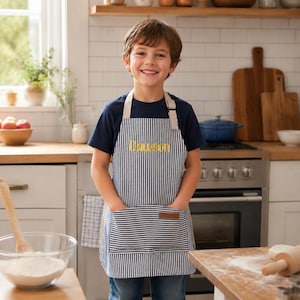 May include: A navy blue and white vertical striped apron featuring the name Dawson embroidered in yellow thread on the chest. The apron includes a large front pocket with a small brown leather-like rectangular patch attached to the top edge.