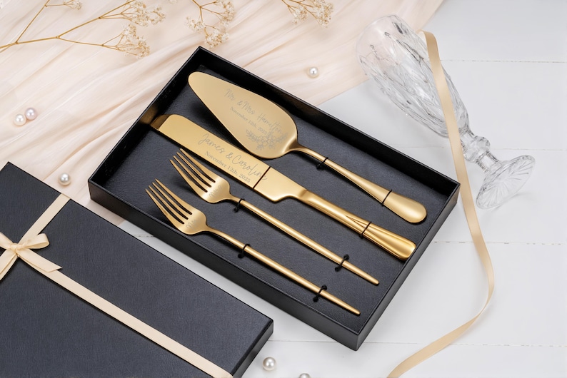 Op de afbeelding: Een goudkleurige bruidstaartservies set, inclusief een serveerschep, mes en twee vorken, gepresenteerd in een zwarte geschenkdoos. De serveerschep en het mes zijn gegraveerd met namen en een datum. Een champagneglas en lint zijn op de achtergrond.
